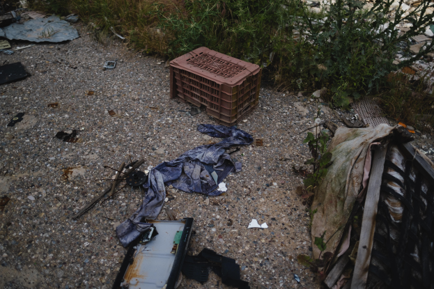 Ein Haufen Müll, darunter ein Plastikbehälter und Kleidung, liegt neben einem Zaun auf einer Müllhalde, mit Pflanzen und Gras im Hintergrund.