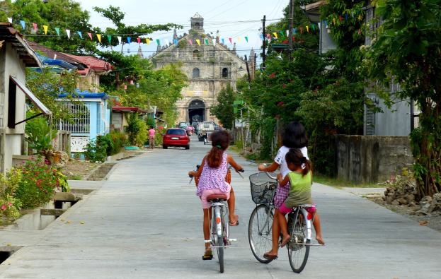 Drei Kinder auf Fahrrädern auf einer von Bäumen gesäumten Straße mit Gebäuden auf beiden Seiten, ein Auto und ein Gebäude im Hintergrund und Himmel oben sichtbar.