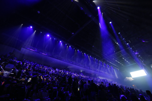 Eine große Menschenmenge sitzt in einer Konzertarena und schaut auf eine Bühne mit Deckenleuchten und einem Bildschirm auf der rechten Seite.