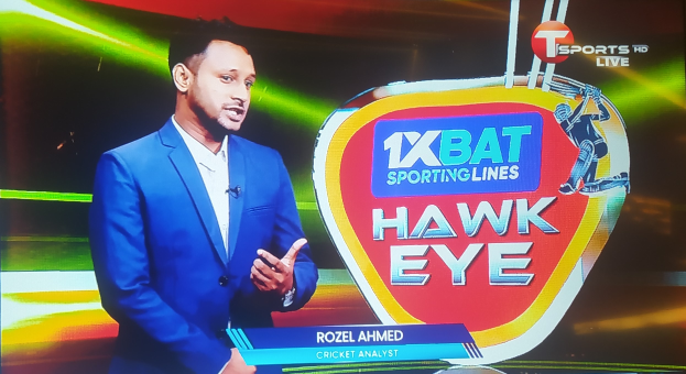 Ein Mann in einem blauen Anzug steht vor einem Fernseher mit einem Logo und Text, der ein Mikrofon tr├Ągt und damit als Cricket-Analyst vermuten l├Ąsst.
