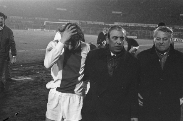 Eine Gruppe verzweifelter Männer in schwarzer und weißer Kleidung steht zusammen auf einem Fußballfeld, mit einem Stadion im Hintergrund, wahrscheinlich nach einem gewonnenen Spiel.