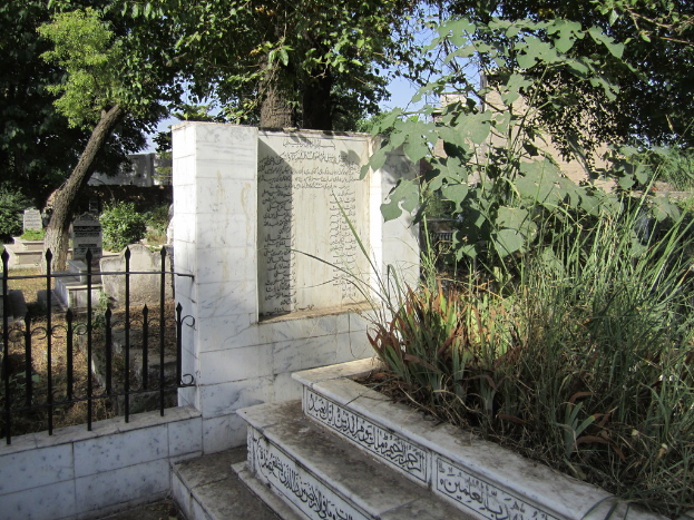 Ein Gedenkschild neben einem Geländer in einem Friedhof, mit Treppen unten, Gras und Bäumen rechts, Gräbern hinter dem Geländer und Gebäuden und Bäumen im Hintergrund.