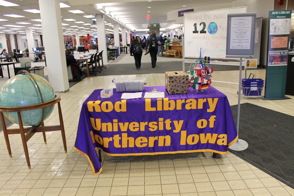 Eine Ausstellung mit einem Tisch, auf dem verschiedene Landesflaggen, eine Box, Papiere und eine Fahne liegen, eine Kugel links daneben, ein Anschlagbrett rechts mit ein paar Leuten in der Nähe und mehreren Tischen und Stühlen links.