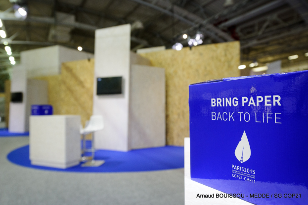 Messestände mit einem blauen und weißen 'Bring Paper Back to Life'-Schild auf der rechten Seite, mit unscharfen Stühlen, Tischen, einem wandmontierten Fernseher und Deckenlampen im Hintergrund.