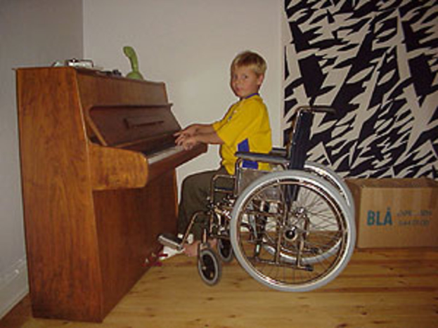 Ein Junge in gelbem Hemd und Hose sitzt in einem Rollstuhl und spielt auf einem Piano mit Spielzeugen darauf, neben einem Vorhang und einer Kiste auf dem Boden.