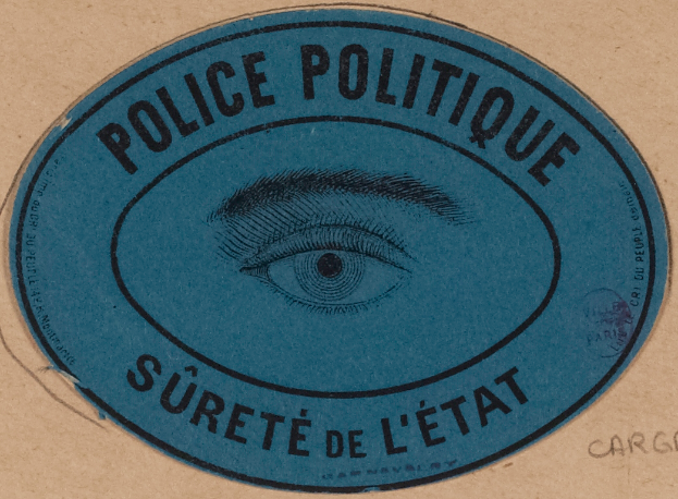 Ein brauner Karton mit einem blauen Aufkleber, auf dem "Polizei Politique Süreté de l√Čtat" steht und ein Auge in der Mitte hat.