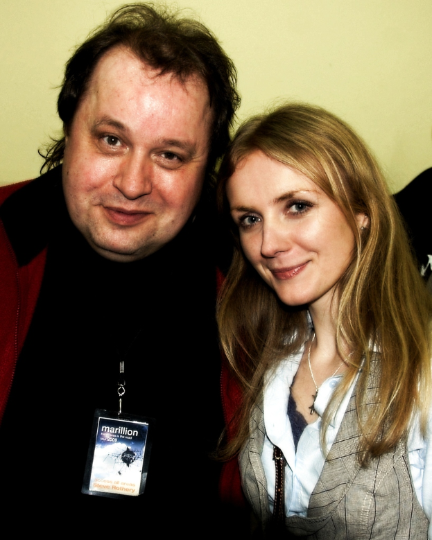 Ein Mann und eine Frau lächeln, mit einem sichtbaren Abzeichen, vor einem hellgelben Hintergrund.