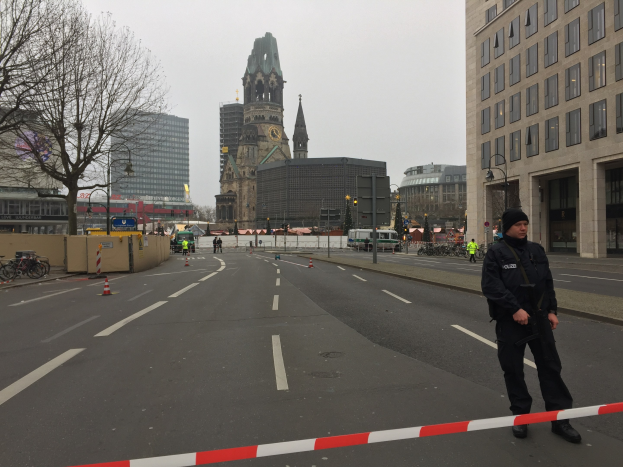 Polizeibeamter in schwarzer Uniform und Mütze auf einer Berliner Straße umgeben von Absperrbaken, Fahrrädern, Laternenmasten, Bäumen und Gebäuden mit einem Kirchturm im Hintergrund unter einem klaren blauen Himmel.