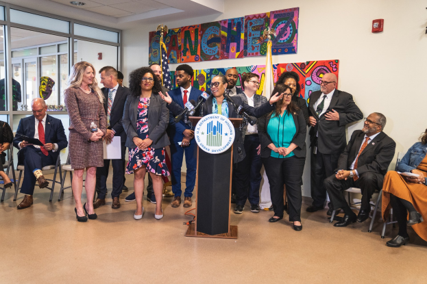 Eine Gruppe von Menschen steht vor einem Rednerpult mit einem Mikrofon und einer Tafel mit Text, während andere auf Stühlen sitzen oder in der Nähe stehen; Flaggen, eine Tafel, ein Feuerlöscher und Deckenleuchten sind an der Rückwand zu sehen.