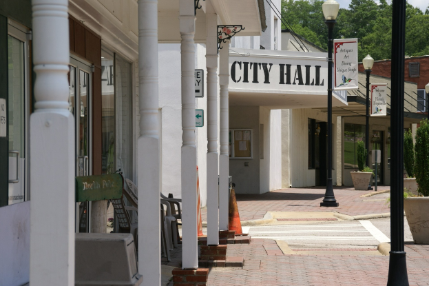 Rathaus mit umliegenden Gebäuden, Straßeninfrastruktur einschließlich Pfosten, Leuchten und Namensschilder, Kraftfahrzeuge, Verkehrskegel, Stühle, Topfpflanzen, Bäume und ein bewölkter Himmel.