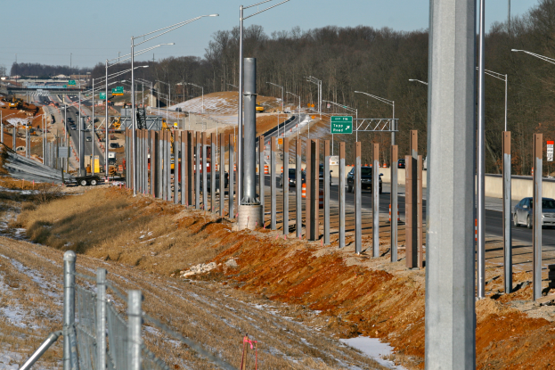 Eine belebte Baustelle mit verschiedenen Fahrzeugen, Pfählen, Lampen und Texttafeln, umgeben von einem Zaun, Gras, Schnee und Bäumen unter einem Himmel.