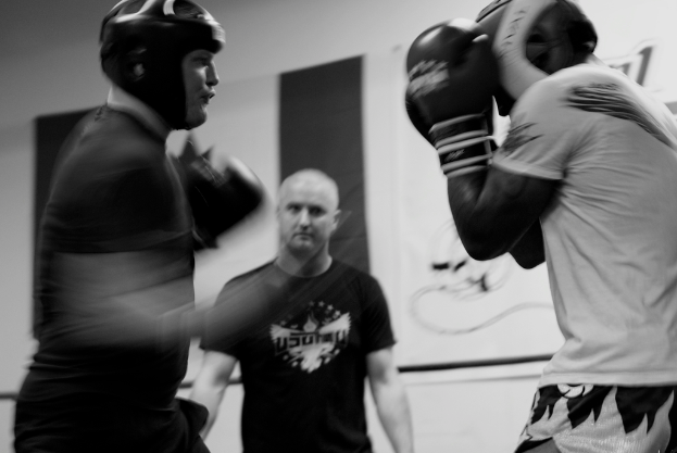 Zwei Boxer mit Helmen und Handschuhen kämpfen im Ring, während ein Schiedsrichter zwischen ihnen steht und ein Banner an der Wand im Hintergrund zu sehen ist.