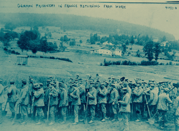 Eine Gruppe deutscher Gefangener in Frankreich mit Waffen im Bild, die auf dem Boden stehen, mit Bäumen, Häusern und einem klaren blauen Himmel im Hintergrund.