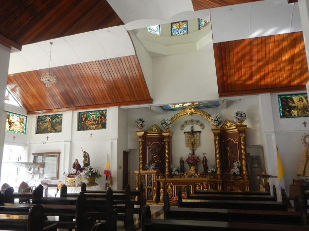 Innere einer Kirche mit hölzernen Kirchenbänken, Statuen, Blumenbouquets, Fahnen, Fotorahmen an den Wänden und Buntglasfenstern, Decke mit Lichtern geschmückt.