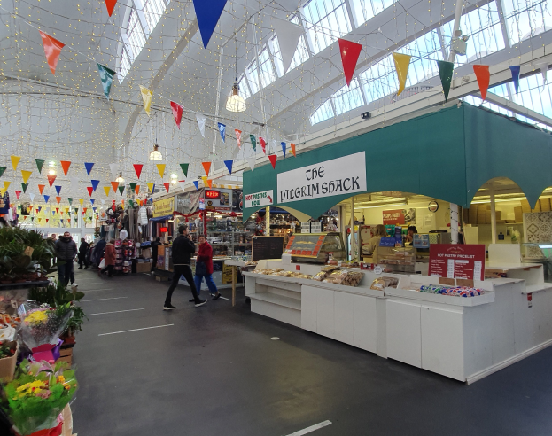 Ein belebter Marktinnenraum mit Blumenbouquets links, Essensständen rechts, einem benannten Stand im Blickfeld und dekorativen Fahnen an der Decke.