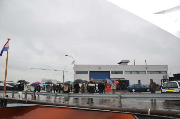 Eine Gruppe von Menschen, die draußen stehen, einige halten Schirme, eine Flagge an einem Mast, zwei Autos, ein Gebäude mit Fenstern, ein Turmkran, eine Straßenlaterne und eine Barrikade.