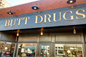 Außensicht eines Gebäudes mit einer leuchtenden Schilder 'butt drugs' und Glaswänden, die einen Drugstore-Interieur mit Fahrzeugen, Gebäuden, Bäumen und Himmel zeigen.