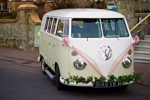 Ein weißer und pinker VW-Bulli, der mit Blumen und Blättern geschmückt ist und vor einer Steinmauer mit einer Treppe auf der rechten Seite und einem Gebäude im Hintergrund geparkt ist.