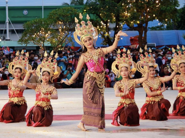 Eine Gruppe von Frauen in traditioneller Thai-Kleidung, die einen Tanz vor einem sitzenden und stehenden Publikum im Freien aufführen, mit Bäumen, Lichtern, Pfosten, einem Gebäude und einem klaren blauen Himmel im Hintergrund.