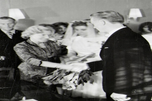 Ein Schwarz-Weiß-Foto eines Brautpaares, das sich die Ringe austauscht, mit der Braut in einem weißen Kleid und Schleier, die einen Blumenstrauß hält, und dem Bräutigam in einem Anzug und Krawatte, mit Wandlampen im Hintergrund.