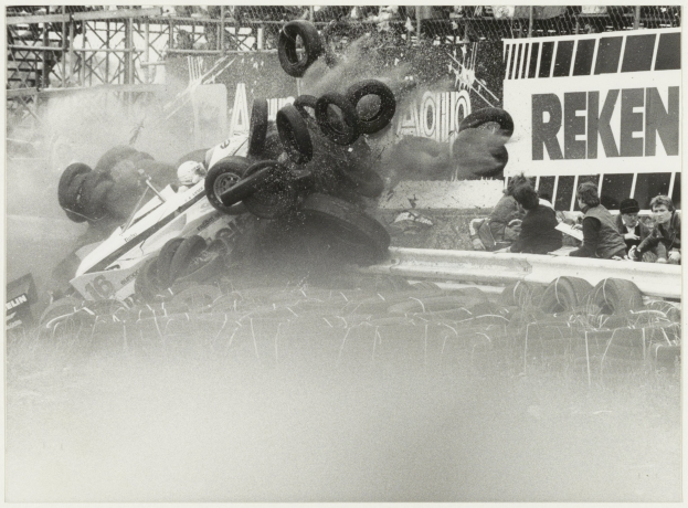 Schwarzes und weißes Foto eines verunglückten Rennwagens, umgeben von Reifen, mit Menschen rechts und einer Tafel mit Text im Hintergrund.
