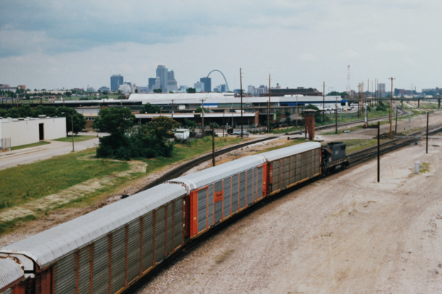 Güterzug auf Schienen neben einer Stadtlandschaft mit Strommasten, Kabeln, Gebäuden, Bäumen, Gras und bewölktem Himmel.