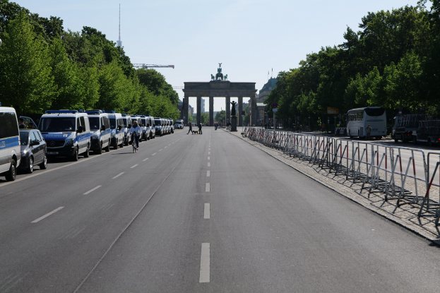 Lange Reihe von Polizeifahrzeugen, die an der Straße vor dem Brandenburger Tor geparkt sind, mit Fahrradfahrern, Fußgängern, Barrieren, Bäumen und einem Bogen mit Statuen im Hintergrund.