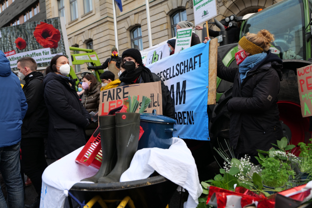 Eine Gruppe von Menschen mit Masken und Protestschildern vor einem Lastwagen, mit einem Tisch voller Gegenstände, Topfpflanzen, Gebäuden und einer Fahne im Hintergrund.