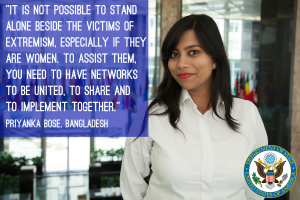 Eine Frau in einem weißen Hemd steht vor einem Gebäude mit einem Zitat über Einheit und Unterstützung für Opfer von Extremismus, insbesondere Frauen. Ein Logo ist in der unteren rechten Ecke sichtbar.