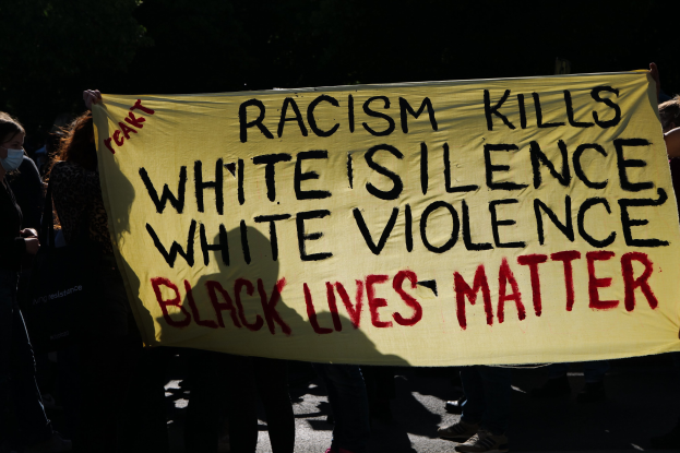 Gruppe von Menschen, die eine "Rassismus tötet, weiße Stille, weiße Gewalt, Black Lives Matter"-Plakate auf einer dunklen Straße halten, alle tragen Masken.