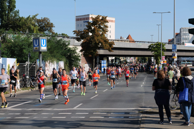 Eine Gruppe von Menschen, die bei einem Marathon auf einer von Bäumen gesäumten Straße mit Laternen, Schildern, Fahrrädern, einem Zaun, Gras, einer Brücke, Gebäuden und einem klaren blauen Himmel laufen.