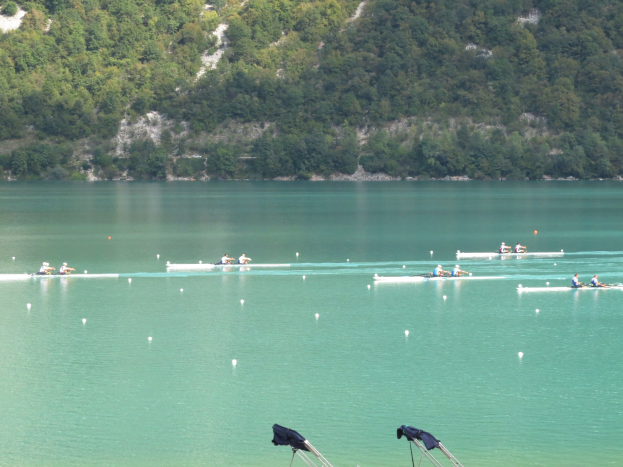 Gruppe von Menschen, die bei einem Wettbewerb auf einem Gewässer rudern, mit Bäumen und einem Hügel im Hintergrund, Kleidung über Stühle am Wasser gelegt.