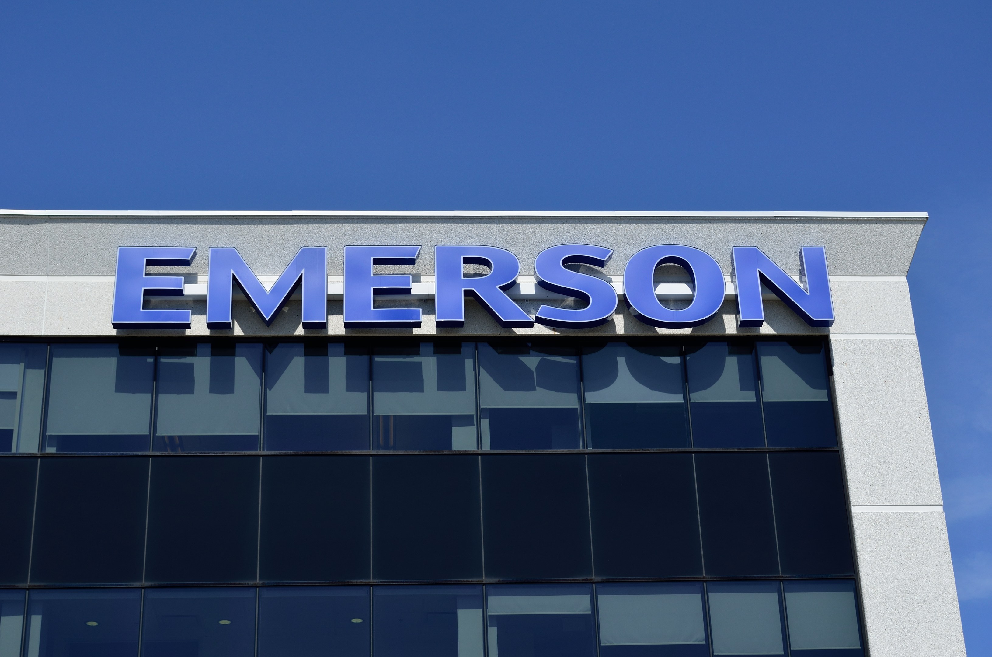 Das Emerson-Logo und der Text "Emerson investiert 1,5 Milliarden US-Dollar in den USA" an der Seite eines Gebäudes vor einem klaren blauen Himmel.