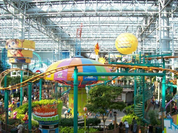Großes Indoor-Wasserpark mit einer Achterbahn, umgeben von Menschen, Bäumen, Pflanzen und Deckenleuchten mit Stangen.