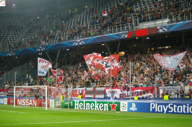 Fußballspiel in einem hell erleuchteten Stadion mit einer großen Menge, grünem Rasen, Tor, Bannern und einem Bildschirm in der oberen linken Ecke.