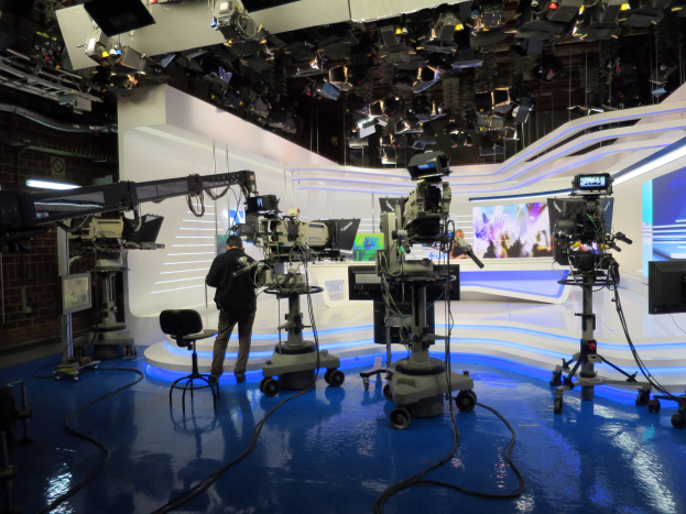 Ein Mann in einer Live-Broadcast-Umgebung, der ein Mikrofon vor einem Fernseher mit Kameras, einem Stuhl, einem Anzeigebildschirm und Deckenleuchten hält.