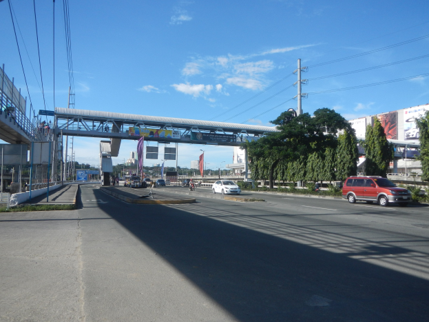 Verkehrsstraße mit Fahrzeugen, Brücke mit Geländern und Pfeilern, Strommasten mit Drähten, Bäume, Gebäude, Banner und ein rotes Auto, das neben der Brücke fährt.