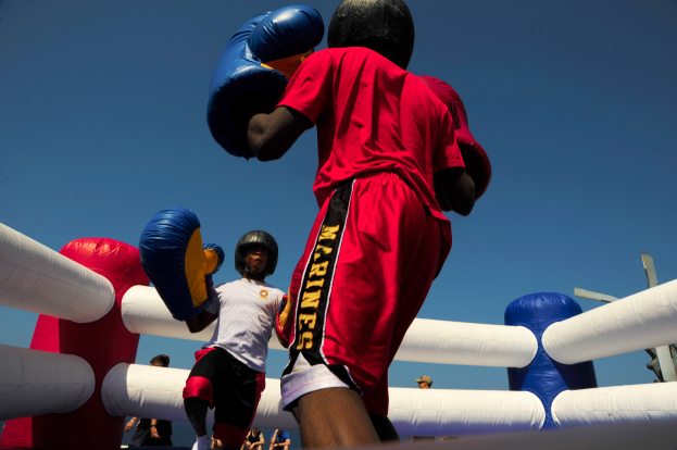 Zwei Boxer im Boxring, einer im roten Shirt und blauen Shorts, kämpfen mit Boxhandschuhen, umgeben von ein paar Leuten und unter einem klaren blauen Himmel.