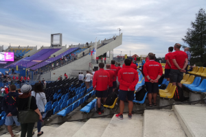 Eine Gruppe von Menschen, einige sitzend und andere auf einer Treppe stehend, versammelt vor einem Stadion mit einem Baum auf der rechten Seite, einem Bildschirm auf der linken Seite, Laternenmasten und einem klaren blauen Himmel im Hintergrund.