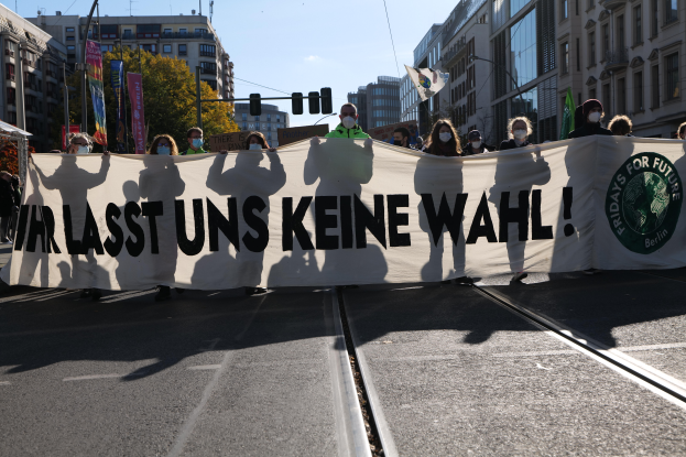 Eine Gruppe von Menschen mit Masken hält ein Banner mit der Aufschrift "Wir sind die letzten keine wahl" vor Gebäuden, Bäumen, Laternenmasten, Ampeln und einem Zelt, mit dem Himmel im Hintergrund.