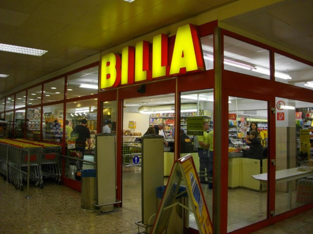 Ein Markt mit einem Eingang in der Mitte, Mülltonnen daneben, einer Reihe von Einkaufswagen auf der linken Seite und einigen Menschen drinnen, die Produkte unter Deckenbeleuchtung durchstöbern.