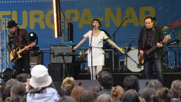 Vier Musiker treten auf der Bühne auf: Eine Frau in einem weißen Kleid singt in der Mitte, flankiert von zwei Gitarristen, mit einem Publikum unten und Lautsprechern an der Wand im Hintergrund.