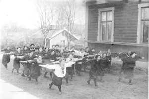 Eine Gruppe von Frauen, die vor einem Haus tanzen, umgeben von Bäumen unter einem klaren Himmel, dargestellt in Schwarz-Weiß.
