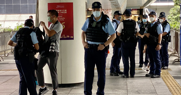 Eine Gruppe von Polizisten in Uniform und Mund-Nasen-Schutz steht vor einem Gebäude, einige halten Kameras, mit einem Banner an einer Säule und Grün im Hintergrund.