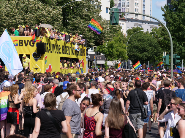 Eine große Menschenmenge, die eine Straße in Berlin, Deutschland, entlanggeht, mit einem gelben Lastwagen im Hintergrund, viele tragen Mützen und halten Fahnen, Laternenpfähle und Verkehrsampeln säumen die Straße, Bäume und Gebäude mit Fenstern im Hintergrund und ein bewölkter Himmel.