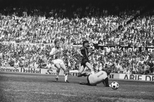 Schwarzes und weiße Foto von Männern, die auf einem Feld Füssball spielen, mit Zuschauern im Hintergrund und einem Banner mit der Aufschrift "Bundesliga 1978-79 Ajax 1-1 Hannover 96."
