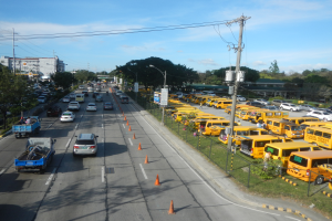 Eine belebte Straßenszene mit verschiedenen Fahrzeugen, Verkehrskegeln, Laternen, Strommasten mit Kabeln und Texttafeln, mit einem Maschendrahtzaun auf der rechten Seite, Bäumen, Gebäuden und einem bewölkten Himmel im Hintergrund.