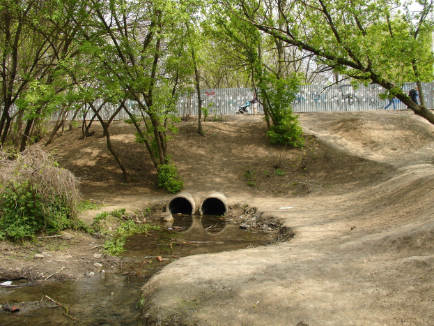Ein Dirtbike-Park mit einem kleinen Bach, umgeben von Bäumen und Pflanzen, mit einer Graffiti-bedeckten Wand im Hintergrund, einigen Menschen und zwei Rohren im Vordergrund unter einem sichtbaren Himmel.