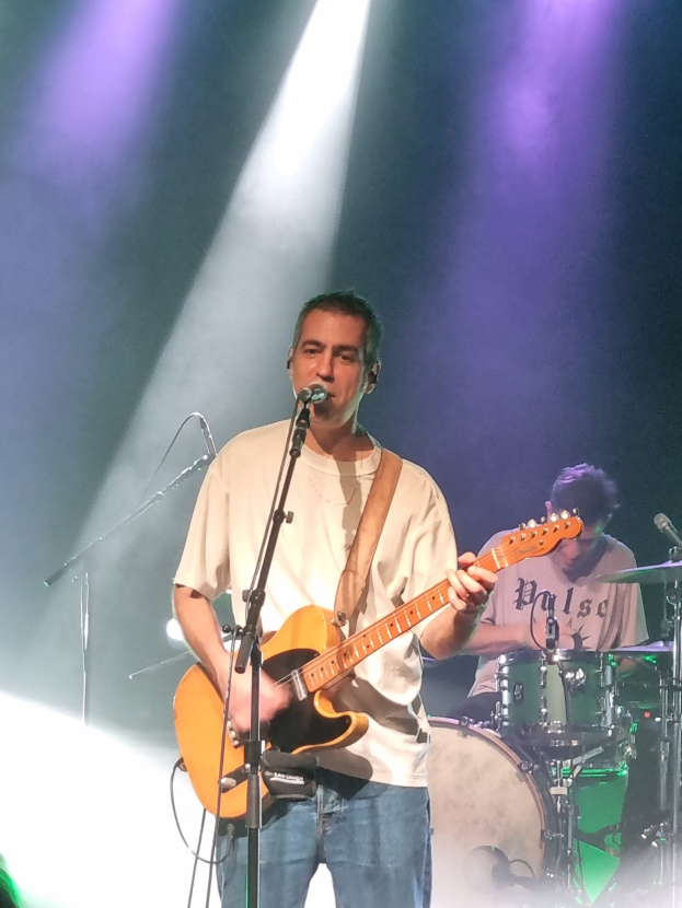 Ein Mann spielt eine E-Gitarre auf der Bühne, beleuchtet von Scheinwerfern von oben, während er in ein Mikrofon singt und die Saiten anschlägt.