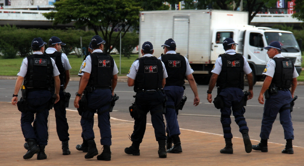 Eine Gruppe von Beamten in Uniform, einschließlich Mützen, Hemden, kugelsicheren Westen, Uhren, Hosen und Schuhen, die auf einer Straße mit einem geparkten Lastwagen auf der rechten Seite neben einem Zaun, Gebäuden, Bäumen, Pflanzen, Pfählen und Bannern im Hintergrund und einer Brücke im oberen rechten Eck gehen.